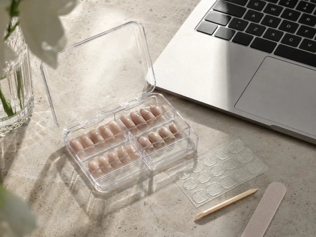 Short neutral press-on nails in a clear plastic case beside a laptop and prep tools
