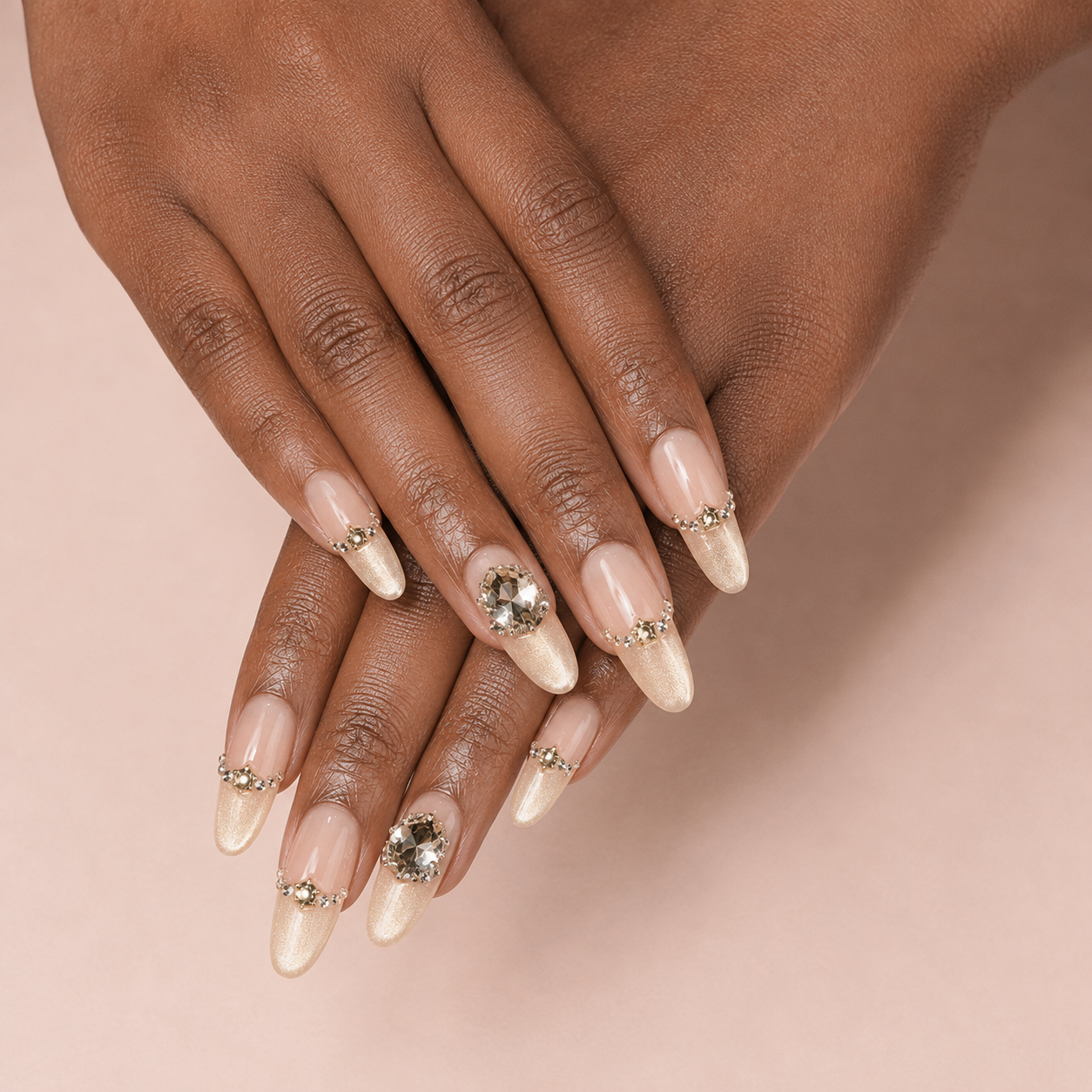 Two crossed medium-brown hands wearing Gilded Cadence Long Oval press-on nails with champagne shimmer and smoky crystals