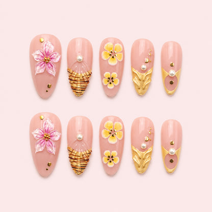 Gilded Hibiscus is a 24-piece handmade medium almond press-on nail set in blush pink, petal pink, yellow and gold, featuring raised flowers, sculptural gold leaves, fan-like gold linework, pearls and studs. It is designed as a reusable multi-size at-home manicure with no selectable size variant, flat lay
